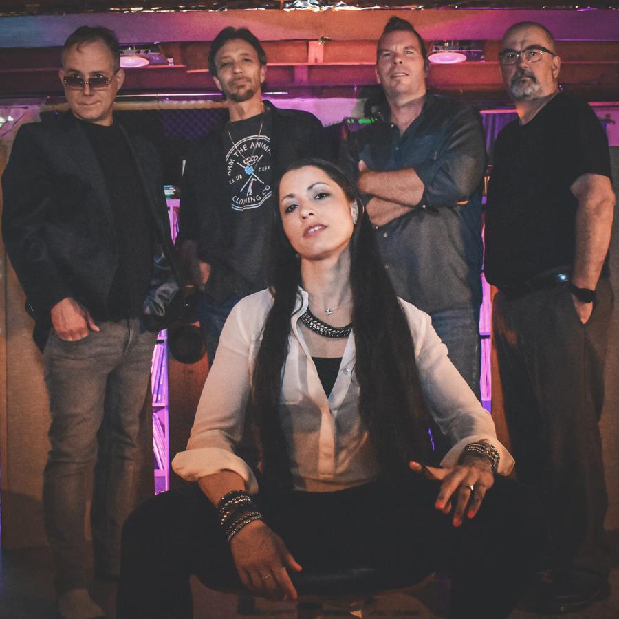 The five members of KG & The Damaged Goods on a venue stage under purple and pink lighting, the vocalist seated centre-front.